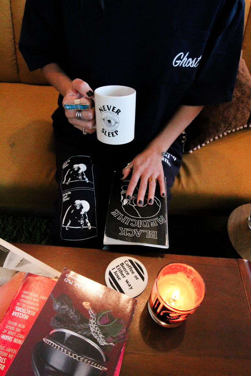 A person sitting on a brown leather couch holds a white mug that reads 'NEVER SLEEP' while surrounded by black graphic merchandise, including a t-shirt with 'Ghost' printed on the chest, a book or album cover titled 'BLACK MAGICIAN', coasters, and a lit candle on a wooden table.
